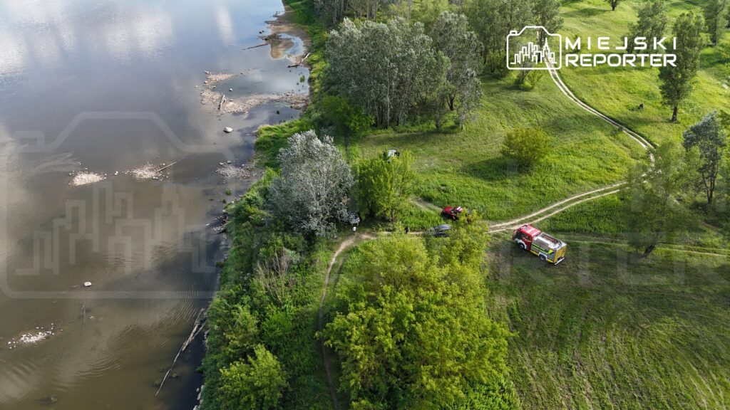 W tle spokojna rzeka, a na brzegu widoczne pojazdy oraz grupka ludzi w otoczeniu zieleni.