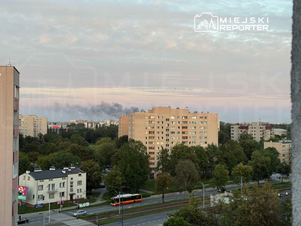 Gęsty, czarny dym unosi się nad zabudowaniami mieszkalnymi w tle, w otoczeniu zielonych drzew i ulicy z tramwajem.