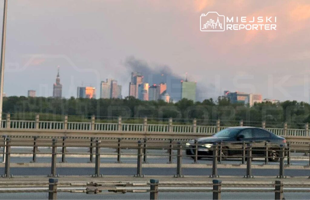 Czarny samochód osobowy przejeżdża po drodze, w tle widać skyline miasta z unoszącym się dymem.