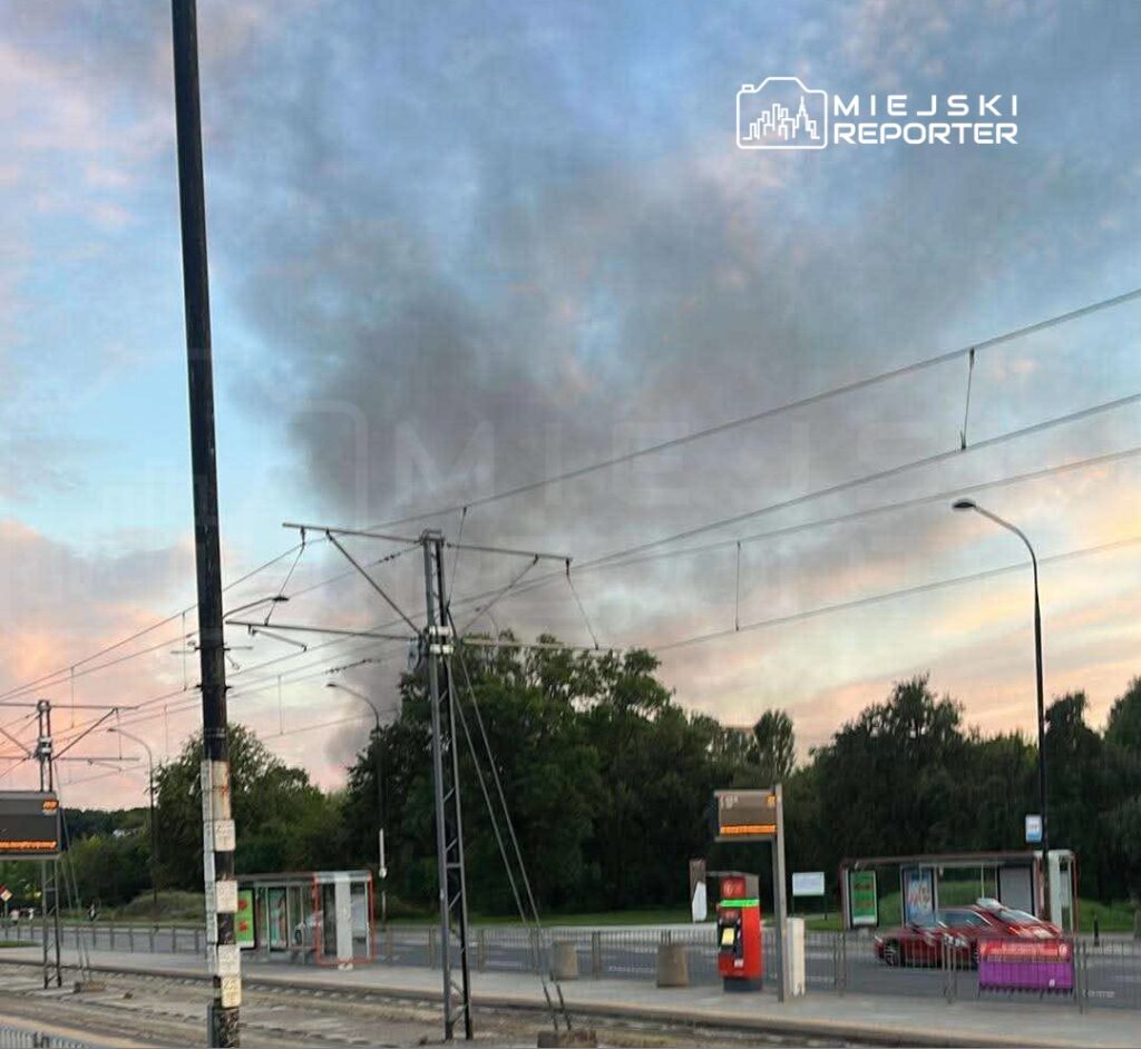 Czarny dym unoszący się nad zadrzewionym terenem w tle, w pobliżu przystanku tramwajowego.