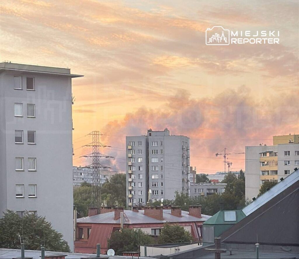 W tle widać szare chmury dymu, a na pierwszym planie nowoczesne budynki mieszkalne oraz dachy domów.