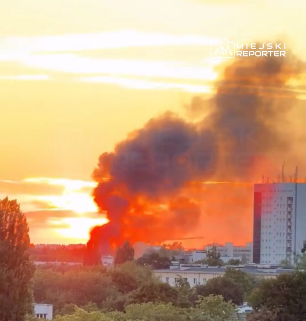 Gęsty, czarny dym unosi się nad horyzontem podczas zachodu słońca, obok nowoczesnych budynków.