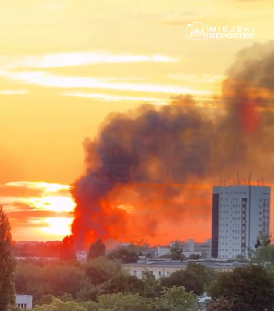 Czarny dym unoszący się w niebo na tle zachodzącego słońca nad miastem. W tle widoczny nowoczesny budynek mieszkalny.