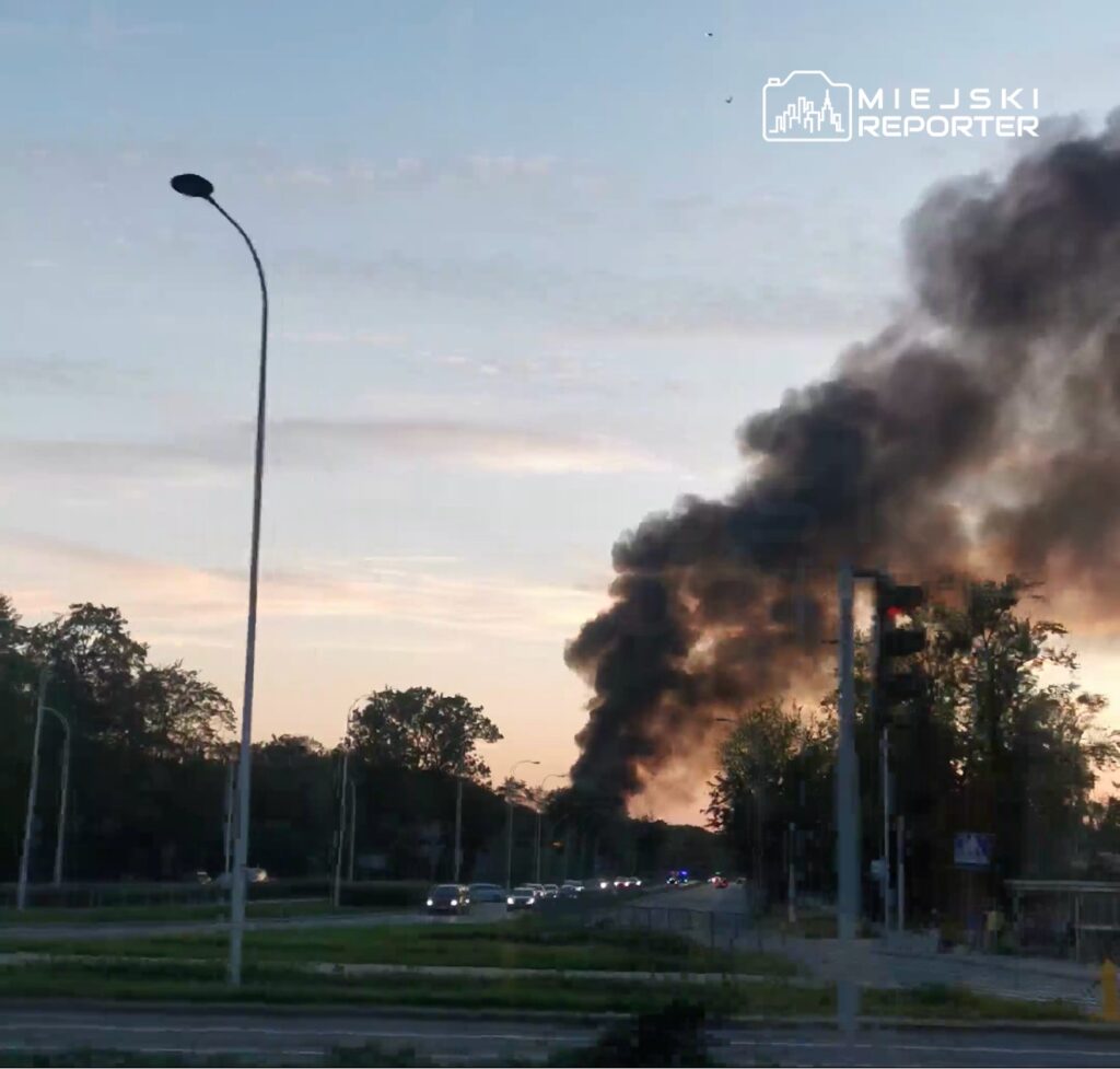 Czarny dym unoszący się nad drogą, w tle zachodzące słońce i samochody poruszające się w kierunku dymu.