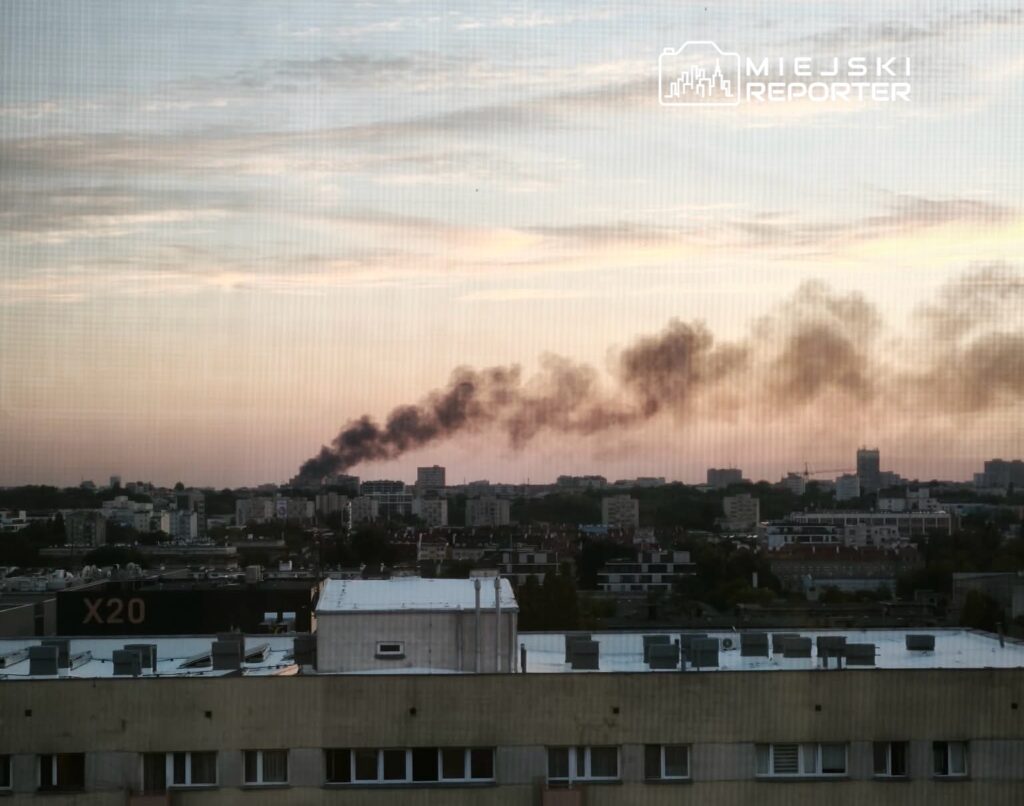 Czarny dym unoszący się nad miastem w tle zachodzącego słońca, widok z okna na zabudowę miejską.