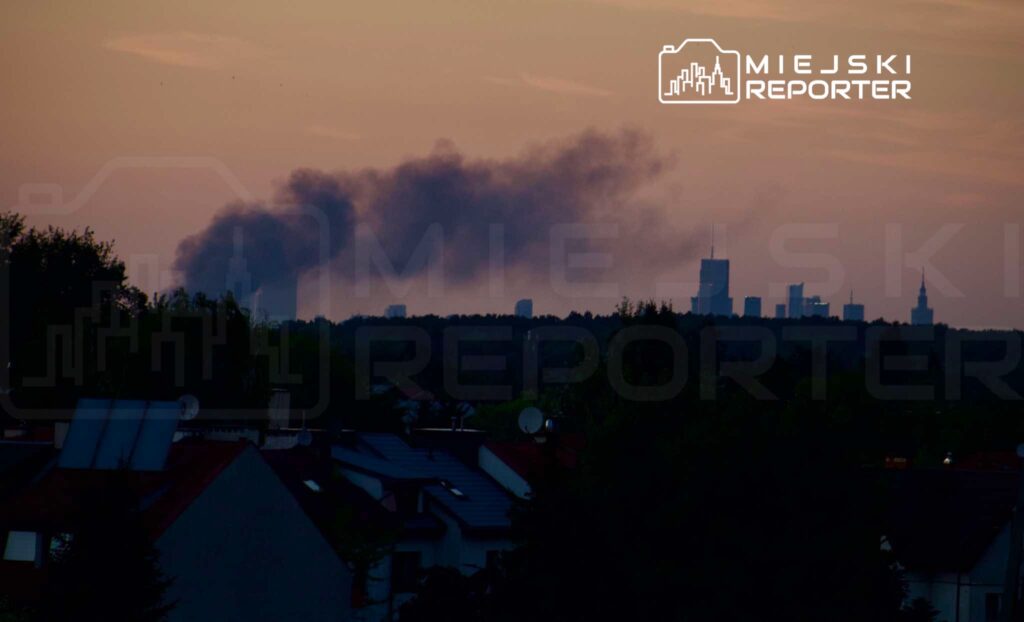 Na tle zachodzącego słońca widać dym unoszący się nad miejskim krajobrazem, w tle wieżowce.