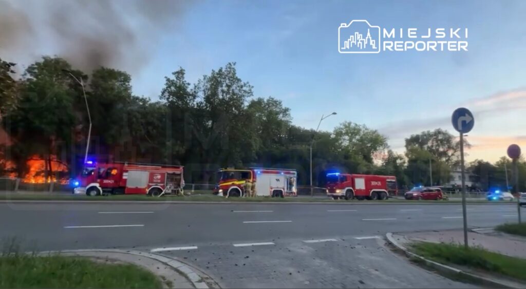 Wielka akcja ratunkowa z udziałem kilku wozów strażackich przy drodze, w tle widoczne płomienie i dym.