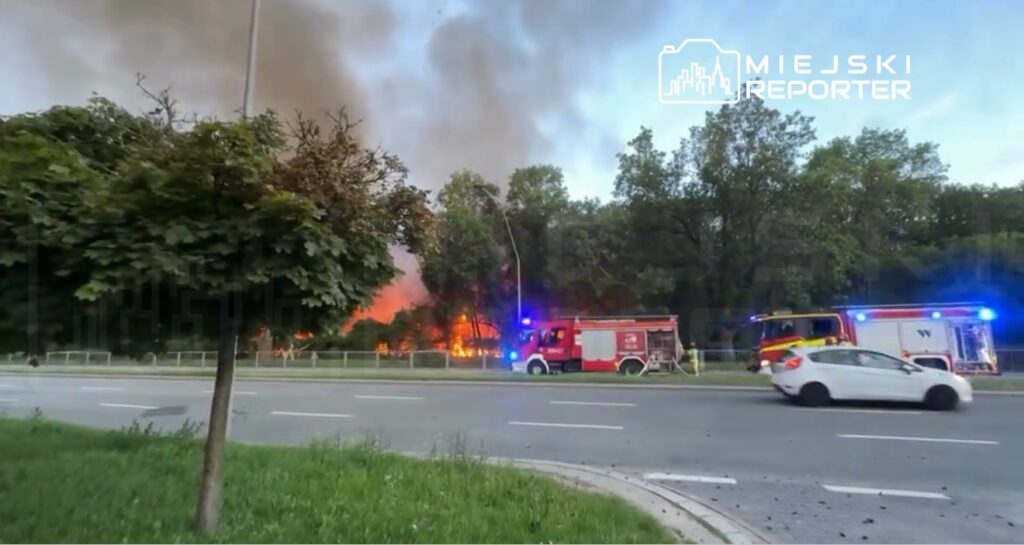 Pojazdy strażackie z włączonymi sygnałami świetlnymi gaszą pożar w lesie, w tle widoczne gęste kłęby dymu.