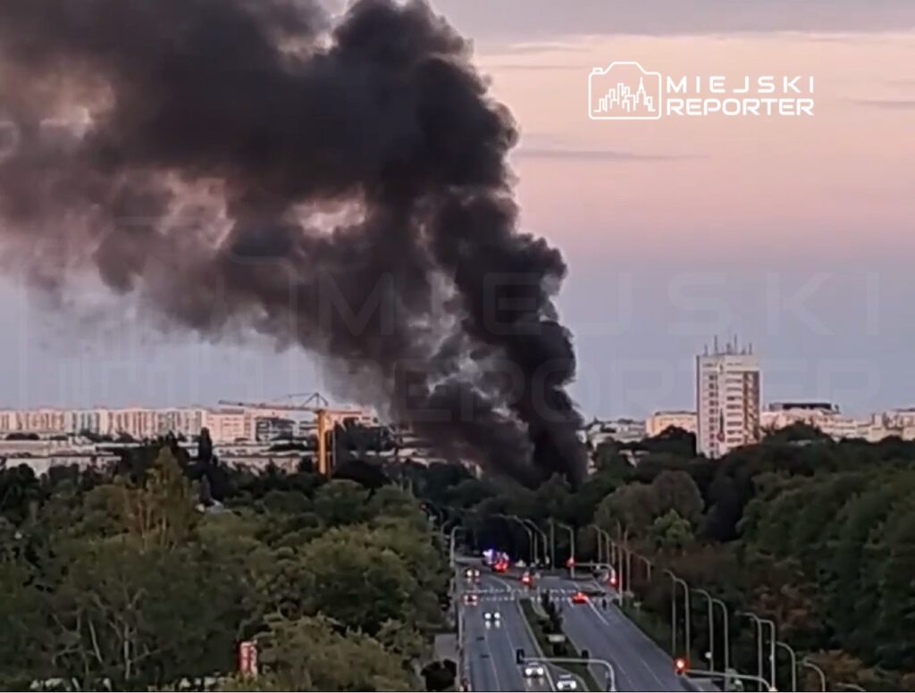 Gęsty, czarny dym unoszący się nad miastem, w tle widoczne budynki mieszkalne i drogi.