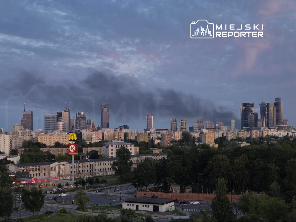 Nowoczesne wieżowce w tle z dymem, w pierwszym planie znak McDonald's oraz stacja benzynowa w miejskim otoczeniu.