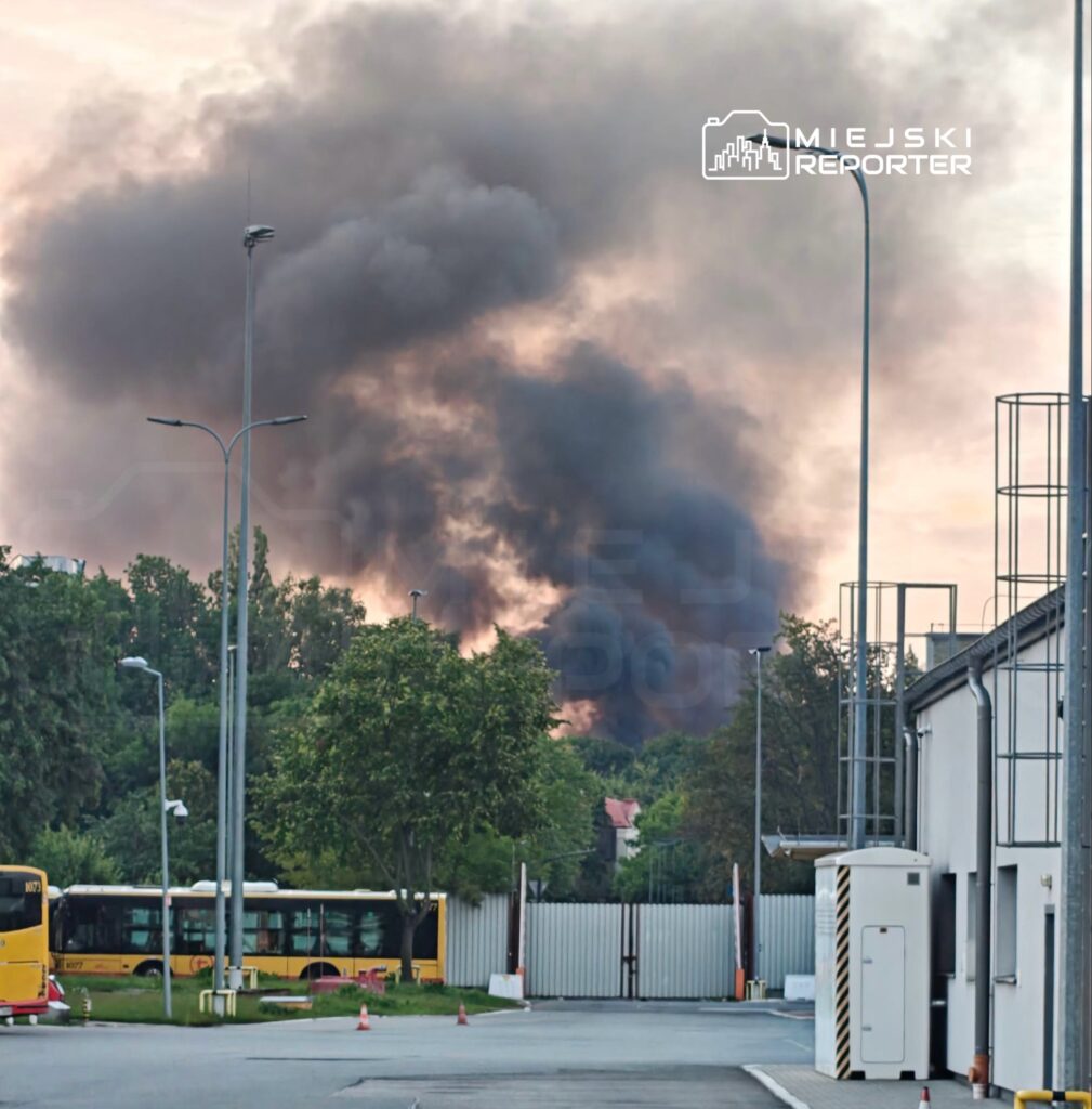 Gęsty, czarny dym unoszący się nad drzewami w tle, w pobliżu miejskiego przystanku autobusowego.