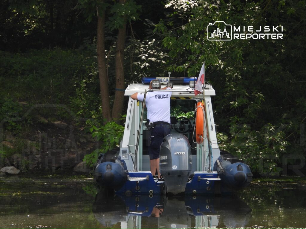 Policjant w mundurze sprawdzający sprzęt na policyjnej łodzi patrolowej przy brzegu zbiornika wodnego.