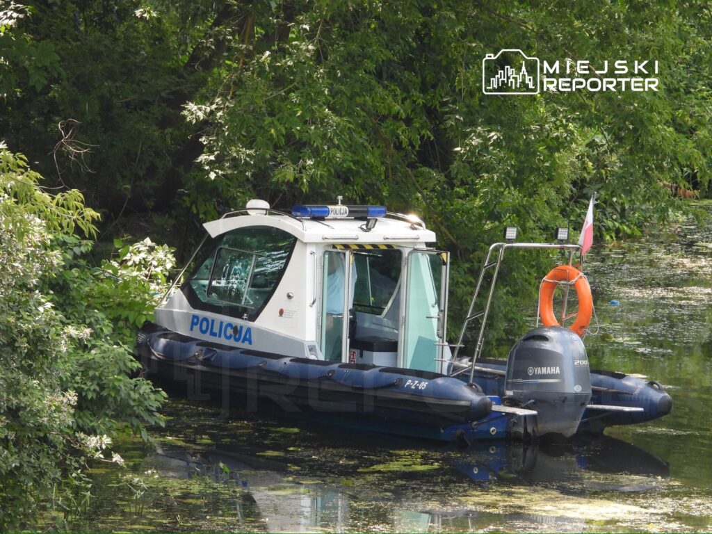 Policyjna łódź patrolowa cumująca przy brzegu otoczona gęstą roślinnością nad wodą.