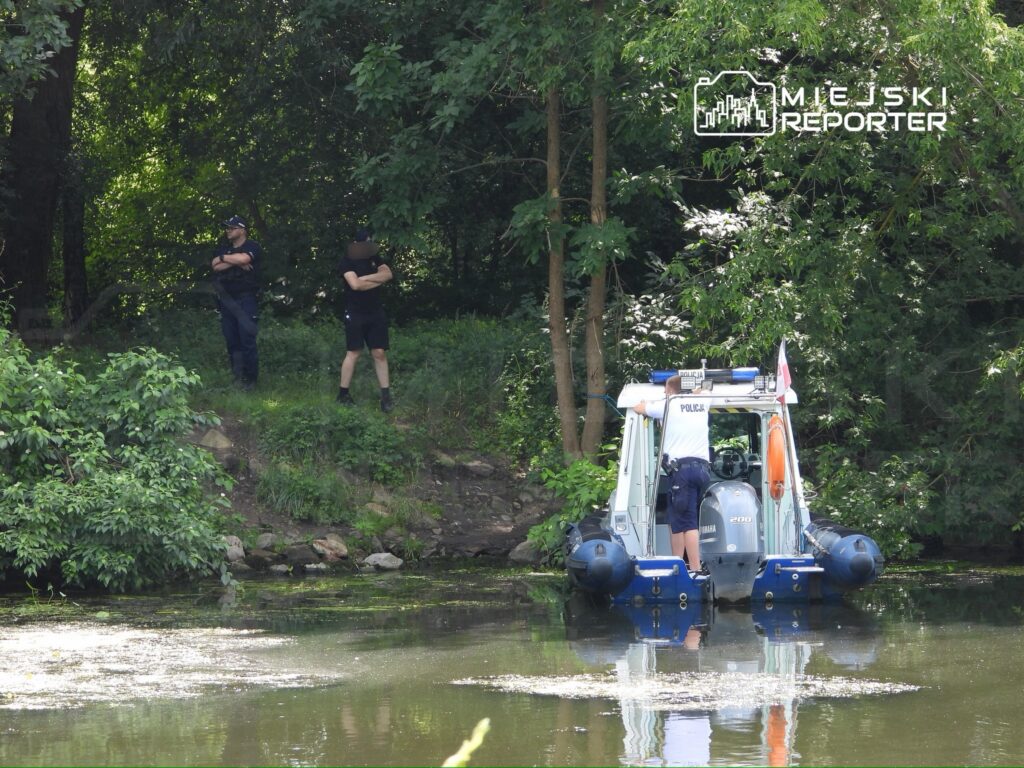 Dwaj policjanci obserwują sytuację z brzegu, podczas gdy trzeci sprawdza wodę z łodzi patrolowej.