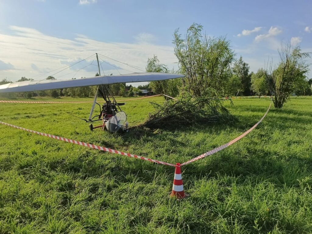 Motolotnia zaparkowana na zielonym terenie, otoczona taśmą ostrzegawczą, w pobliżu drzew i krzewów.