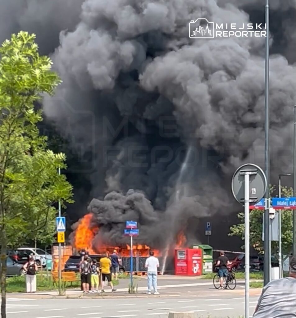 Wielkie płomienie i gęsty dym unoszą się z palących się odpadów przy ulicy, a gapie obserwują zdarzenie z bezpiecznej odległości.