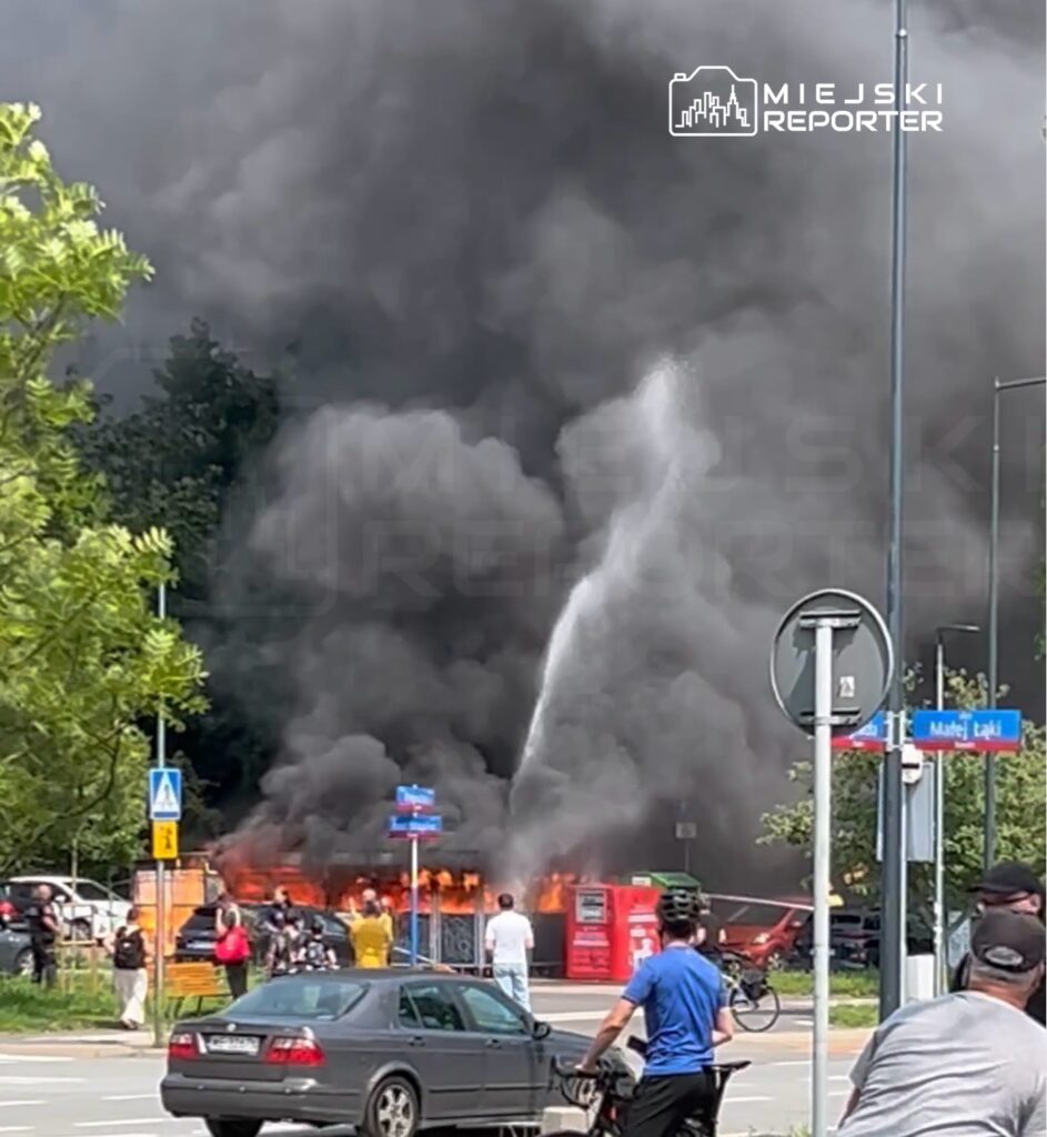 Ogień trawi budynek, a strażacy walczą z płomieniami, otoczeni przez gapiów i dym unoszący się w powietrzu.