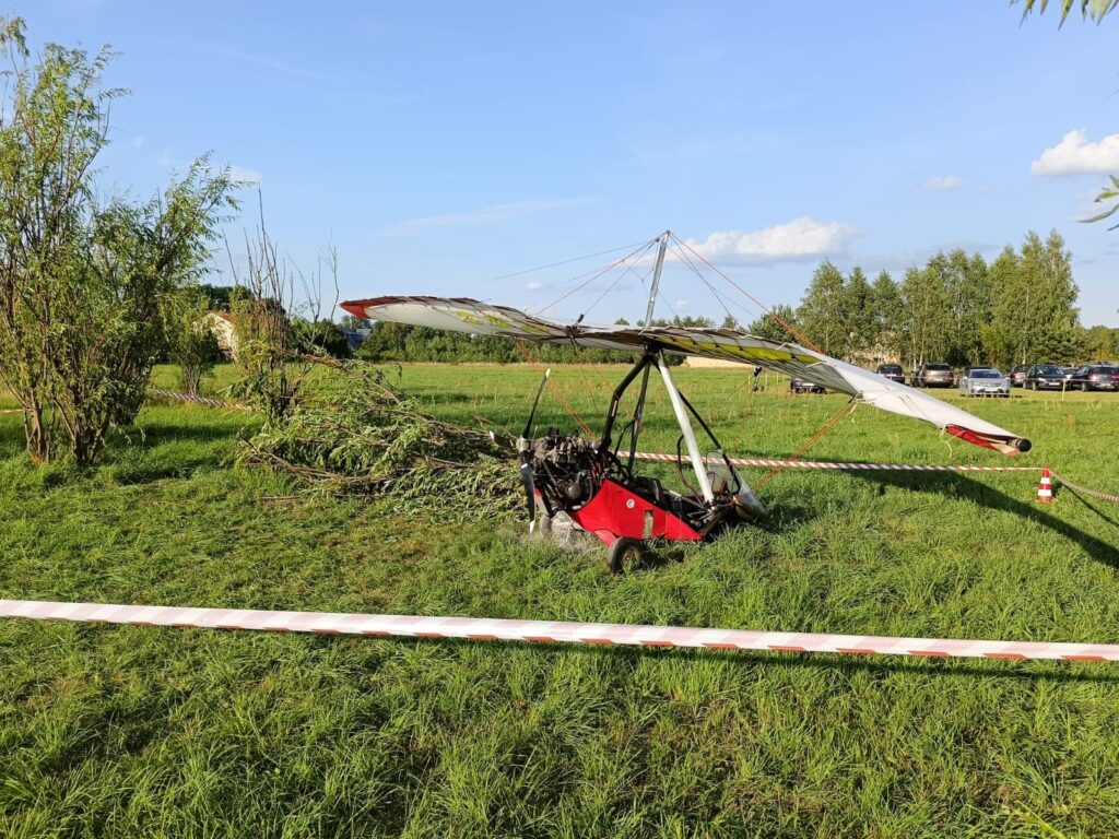 Zniszczony ultralekki samolot leży na trawie wśród drzew, otoczony taśmą ostrzegawczą. W tle widoczne zaparkowane samochody.