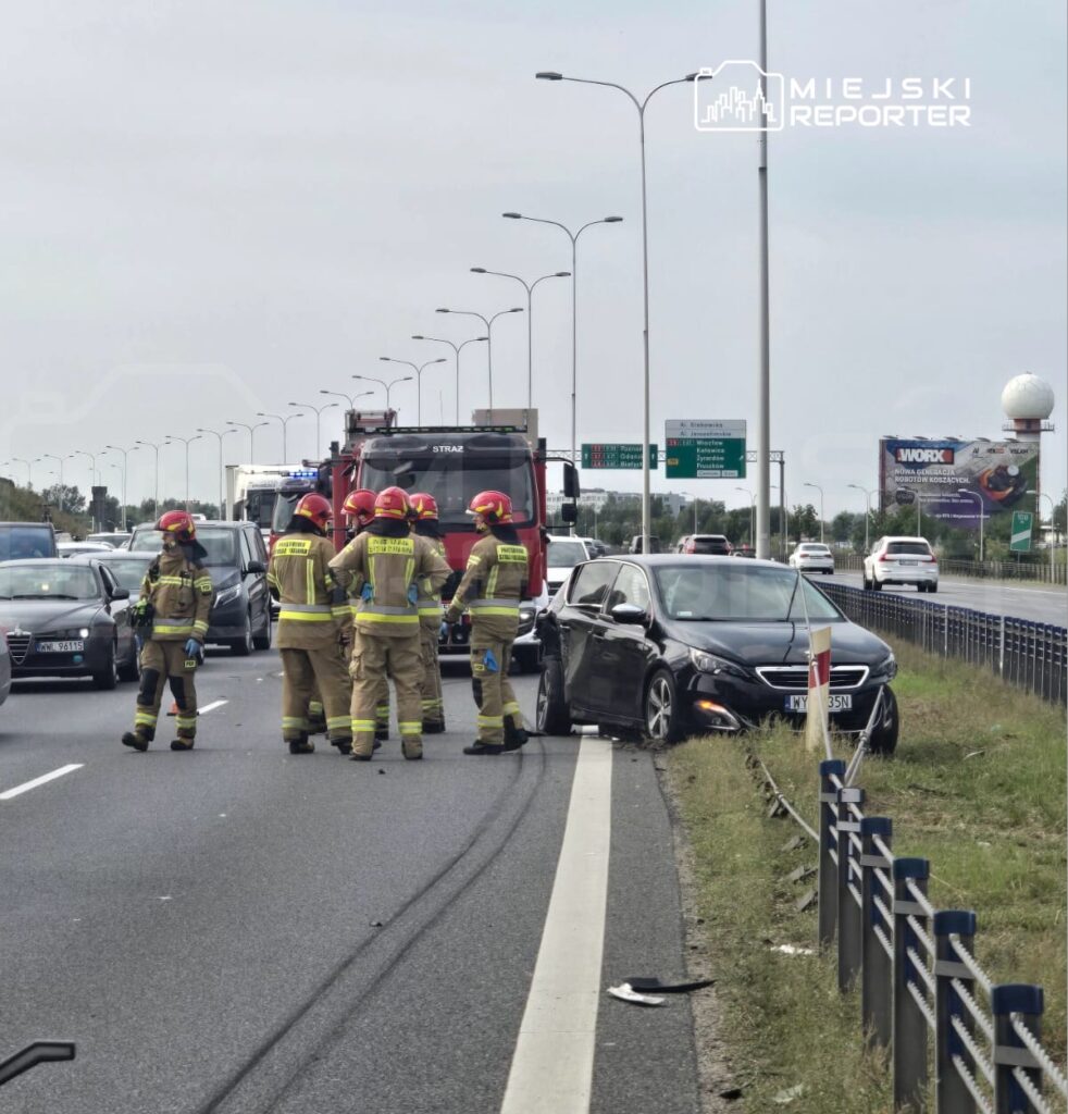 Grupa strażaków w hełmach bada uszkodzony samochód na ruchliwej drodze, obok stoi wóz strażacki.
