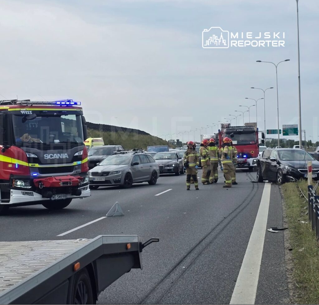 Grupa strażaków w mundurach na drodze, obok uszkodzonego samochodu, przy zablokowanym ruchu na autostradzie.