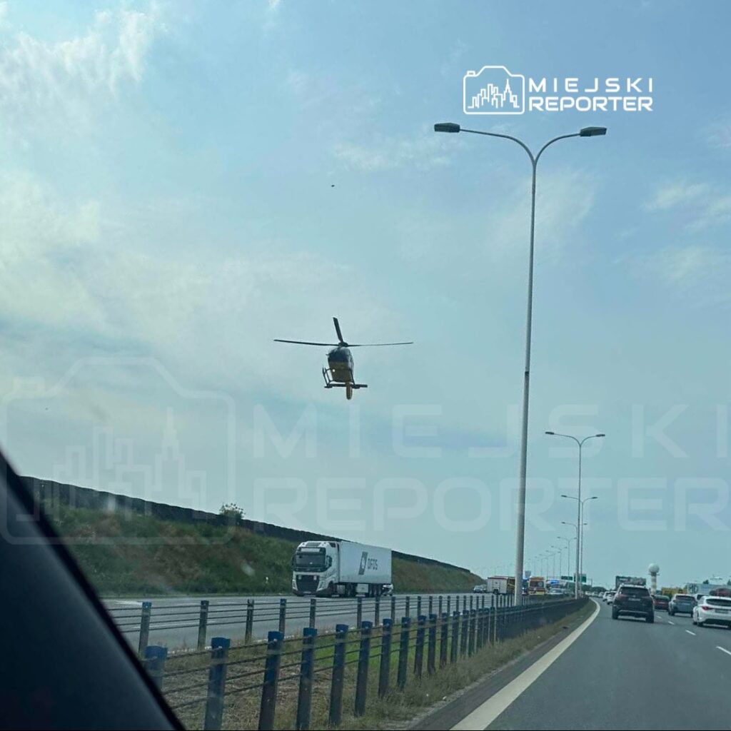 Helikopter przelatujący nad ruchliwą drogą, w tle widoczne samochody i słupy oświetleniowe.