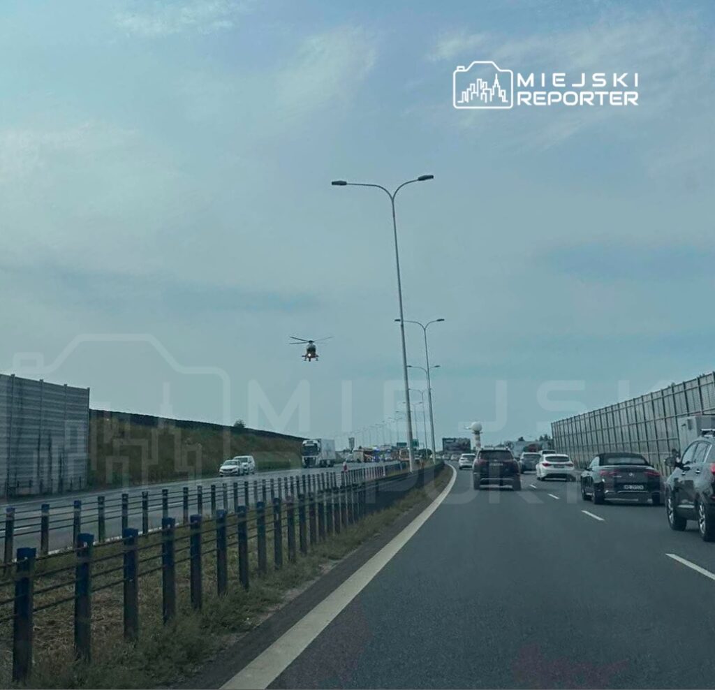 Helikopter unosi się nad ruchliwą autostradą, nad pojazdami w kierunku przeciwnym, w tle widoczne są bariery dźwiękochłonne.