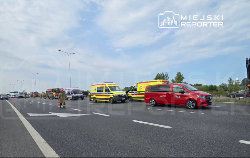 Na drodze stoją pojazdy ratunkowe, w tym karetki i samochód straży pożarnej, w trakcie akcji ratunkowej.