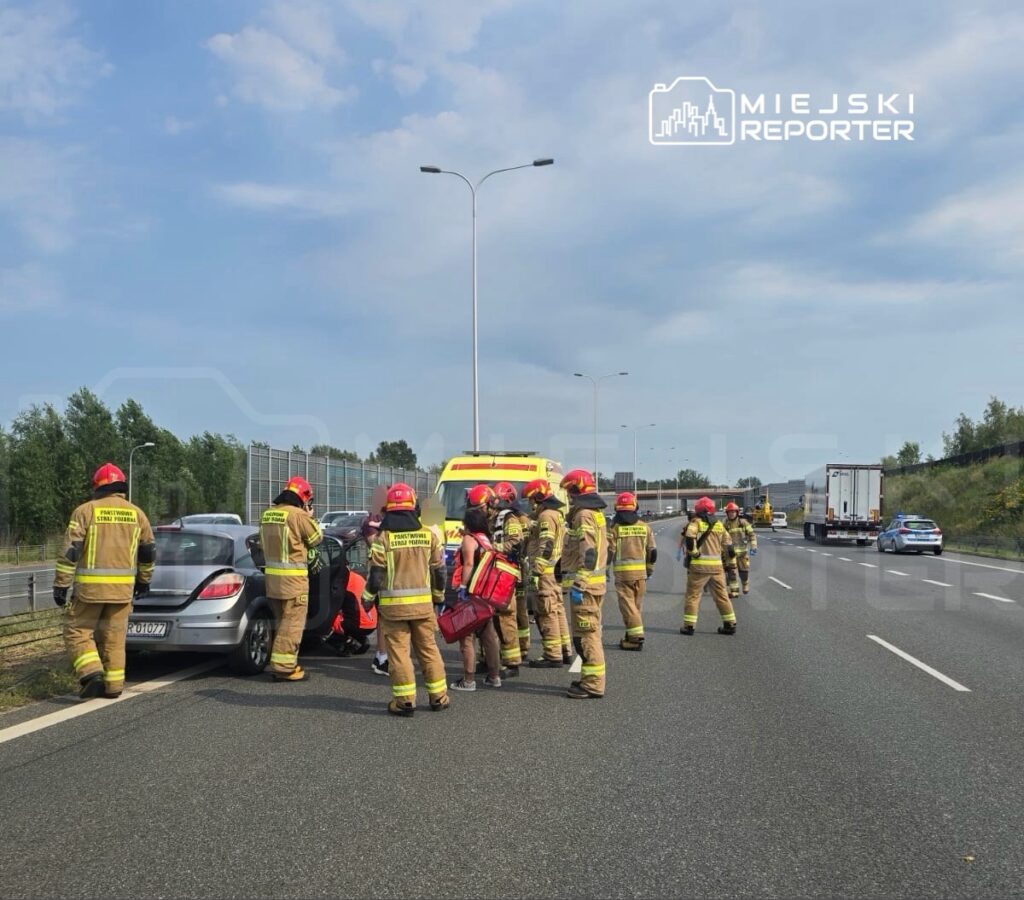 Grupa strażaków w mundurach, z hełmami, udziela pomocy poszkodowanej osobie w samochodzie na drodze.