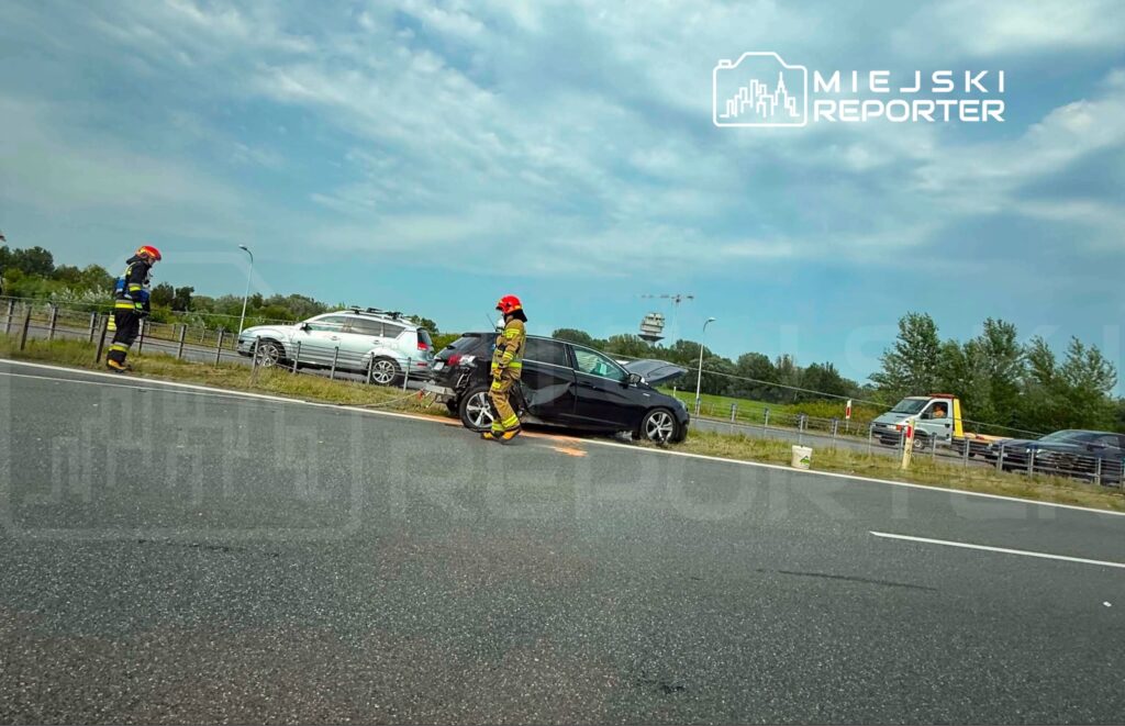 Strażak w mundurze z kaskiem bada uszkodzone czarne auto na poboczu drogi, obok przejeżdżają inne pojazdy.