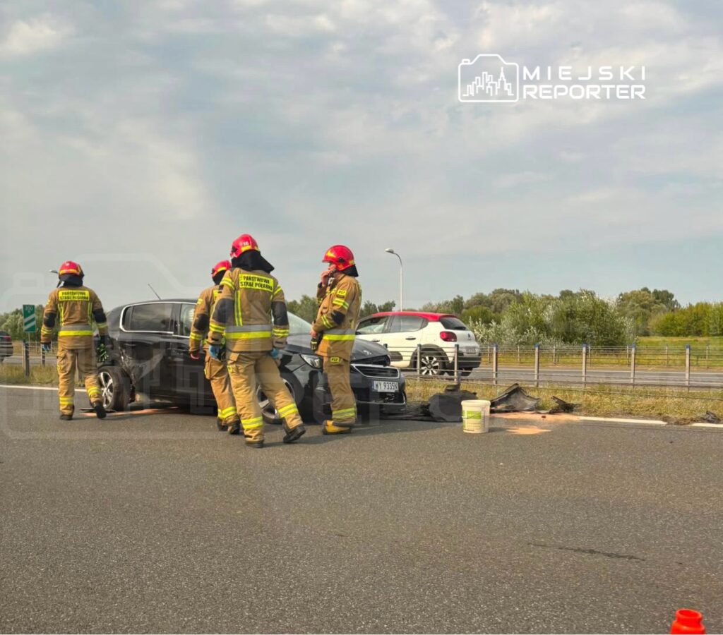 Grupa strażaków w mundurach omawia sytuację przy uszkodzonym samochodzie na drodze.