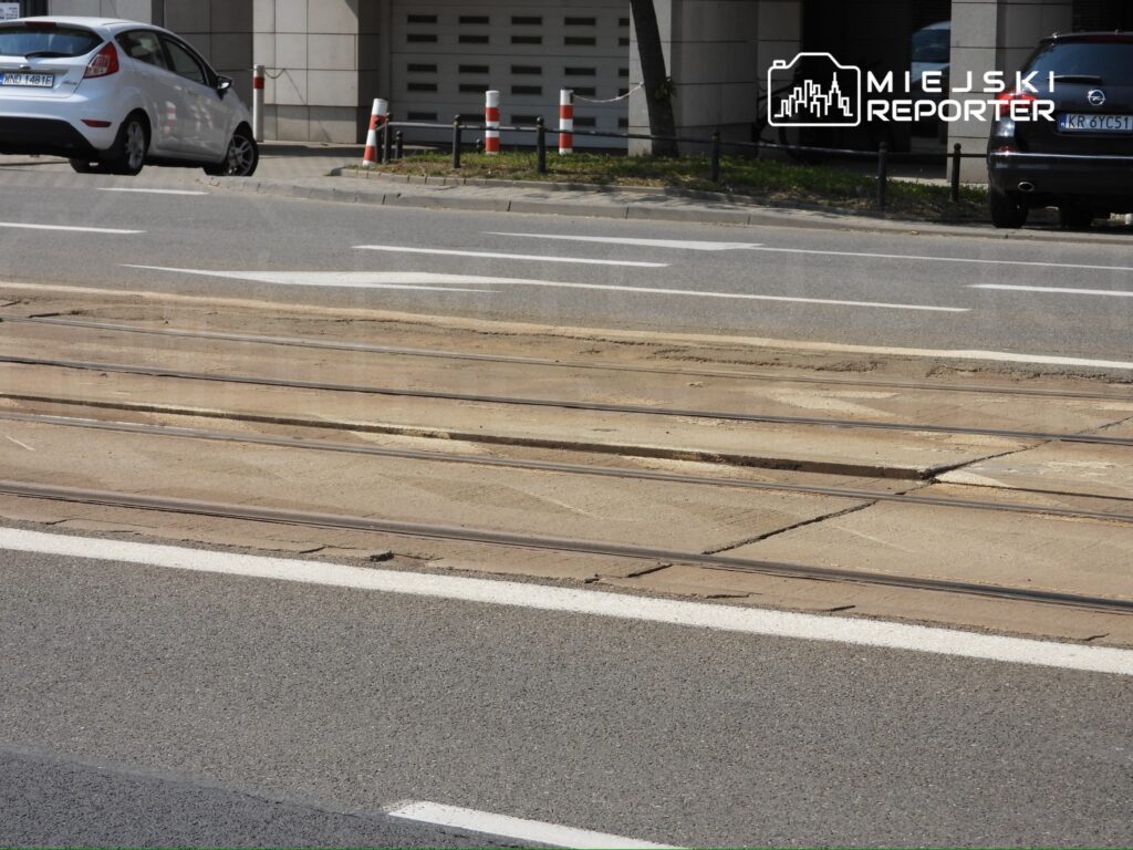 Pusta ulica z widocznymi torami tramwajowymi obok asfaltowej jezdni, przy której zaparkowane są samochody.