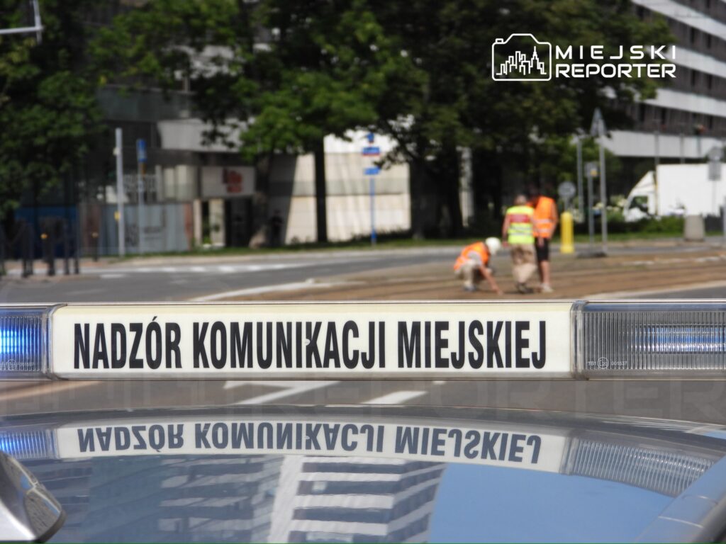 Na pierwszym planie widoczny jest napis "NADZÓR KOMUNIKACJI MIEJSKIEJ" na radiowozie, w tle pracownicy w kamizelkach odblaskowych sprzątają teren.