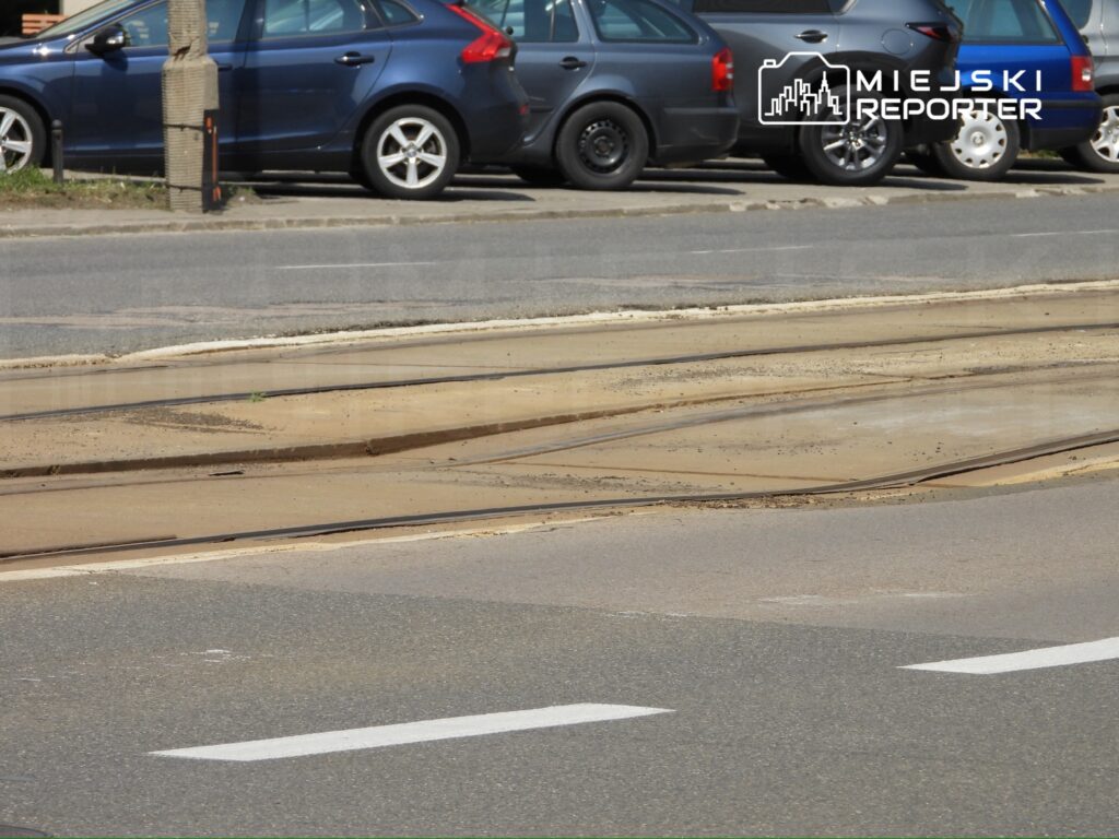 Tramwajowe tory przecinające asfaltową drogę w otoczeniu zaparkowanych samochodów.