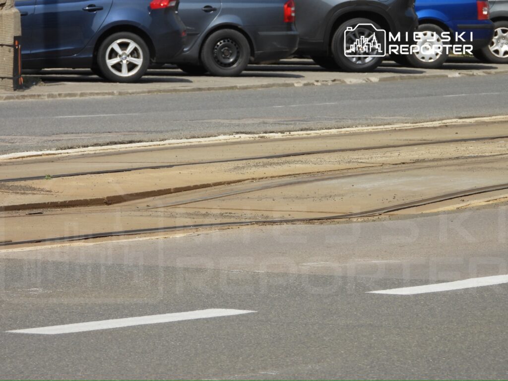 Na parkingu widoczne są samochody stojące obok torów tramwajowych, z wyraźnym podziałem nawierzchni.