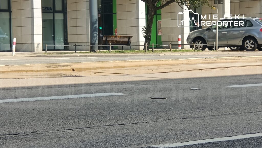 Na ulicy widoczne są znaki drogowe oraz pasy ruchu, obok których parkują samochody. W tle znajduje się nowoczesny budynek z dużymi oknami.