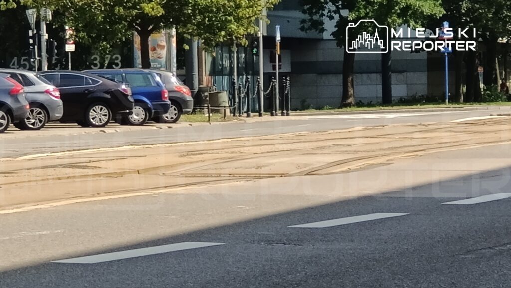 Ruchliwa ulica z zaparkowanymi samochodami oraz widocznymi torami tramwajowymi na nawierzchni.
