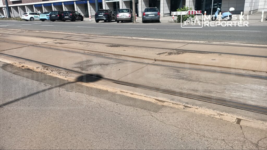 Ulica z torami tramwajowymi, obok zaparkowane samochody, w tle nowoczesny budynek z przeszkloną fasadą.