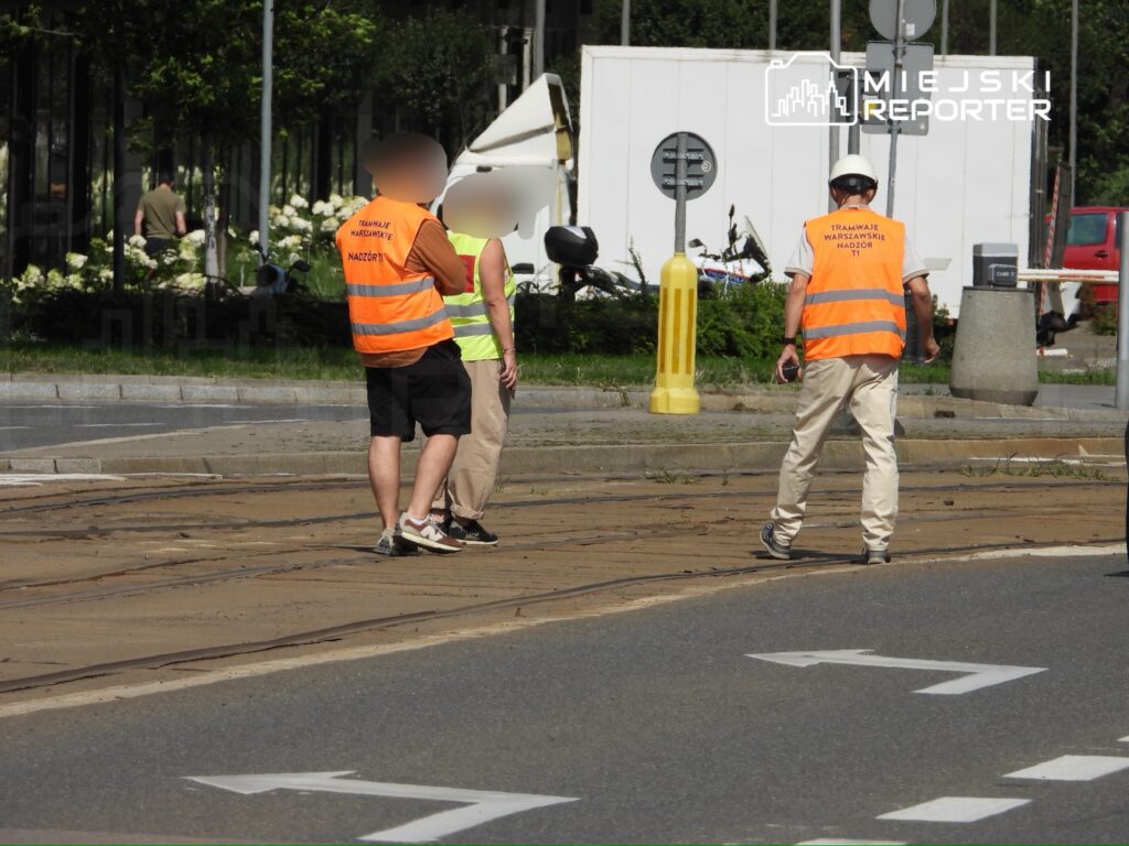 Trzech pracowników w kamizelkach odblaskowych rozmawia na torowisku tramwajowym w słoneczny dzień.