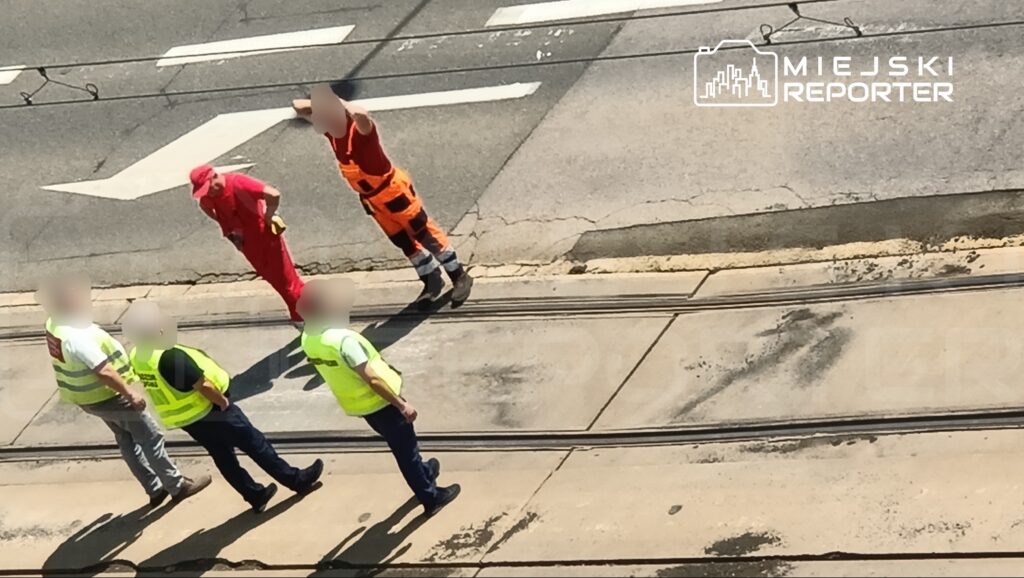 Grupa pracowników w kamizelkach odblaskowych rozmawia z mężczyzną w czerwonym stroju, stojąc na ulicy.