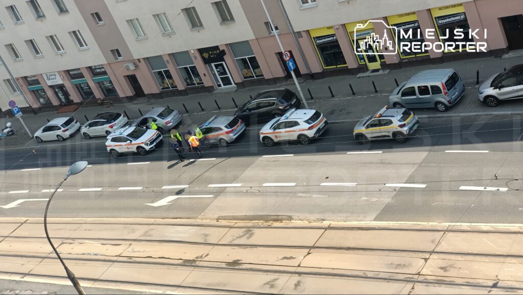 Grupa policjantów rozmawia z kierowcą przy zaparkowanych samochodach na ulicy w mieście.