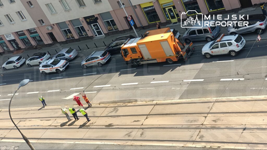 Czterech pracowników w kamizelkach odblaskowych prowadzi prace przy torowisku tramwajowym na ruchliwej ulicy.