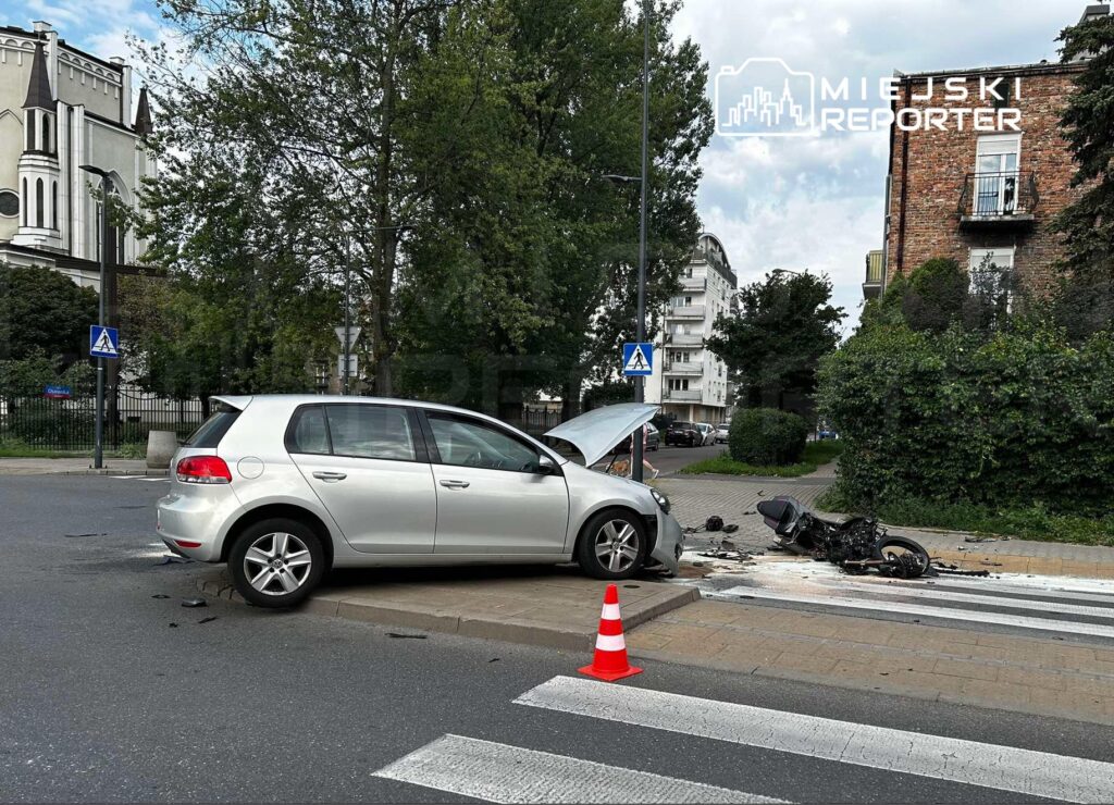 Uszkodzony samochód osobowy z otwartą maską stoi na krawężniku, obok leży rozbity motocykl, a w tle widać zieleń drzew.