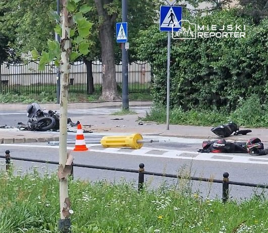 Motocykl leży na jezdni obok przewróconego słupka oraz pomarańczowego pachołka, w tle znaki drogowe.