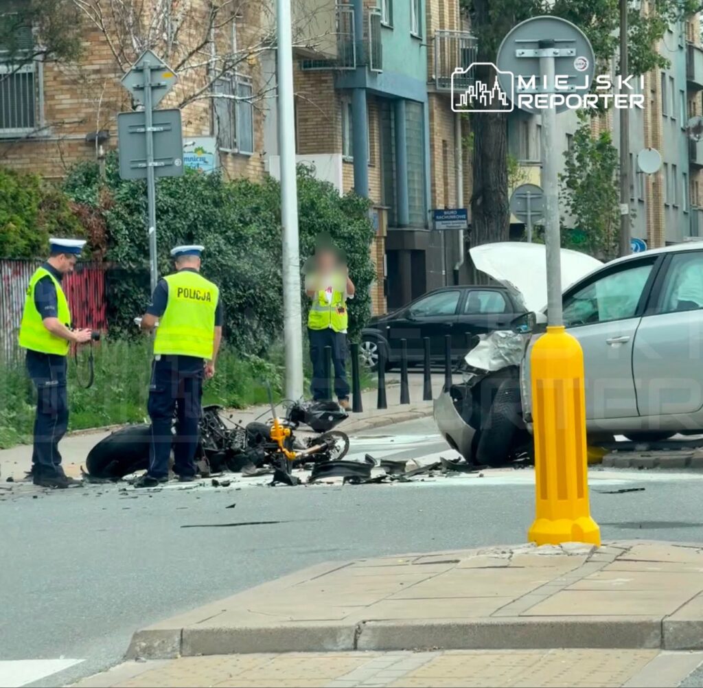 Policjanci w kamizelkach odblaskowych badają miejsce wypadku, gdzie leży uszkodzony motocykl obok rozbitego samochodu.