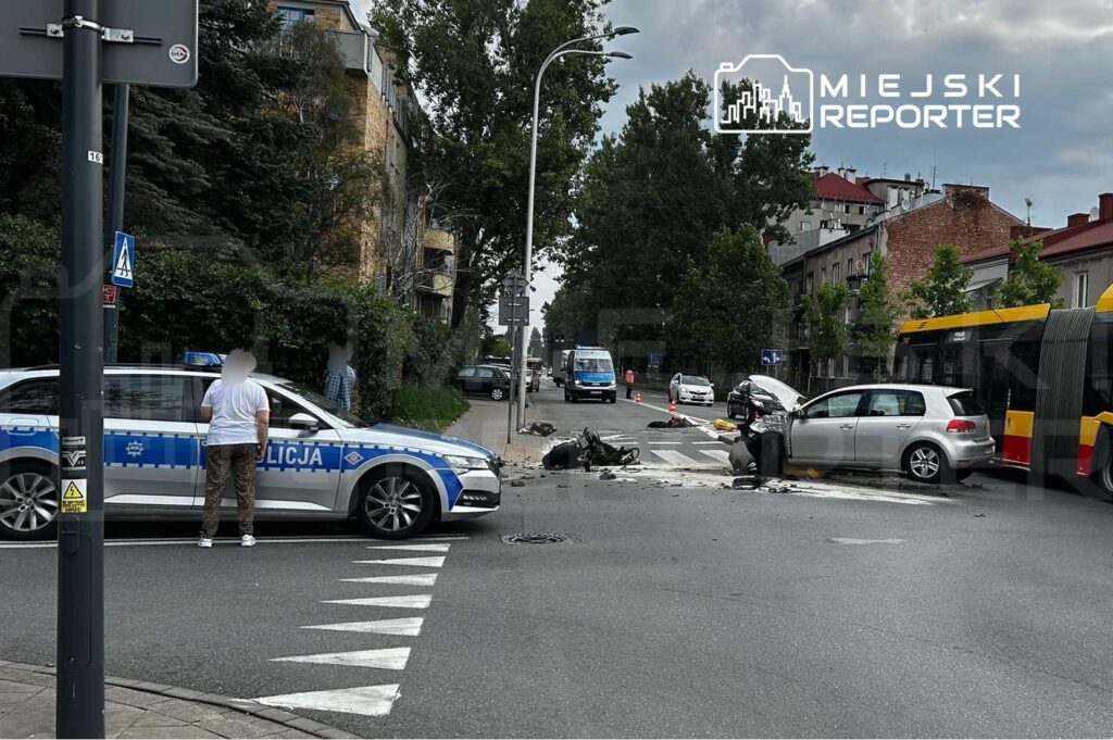 Na skrzyżowaniu ulic stoi radiowóz policji, obok którego leży rozbity skuter i uszkodzony samochód osobowy.