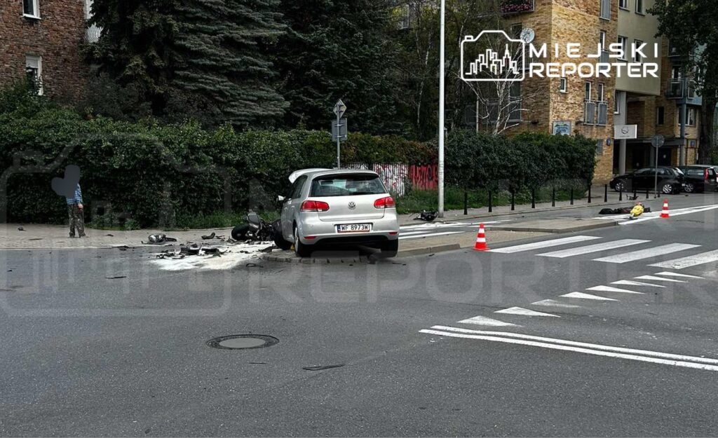 W miejscu wypadku drogowym leży rozbity motocykl obok uszkodzonego samochodu osobowego na chodniku.