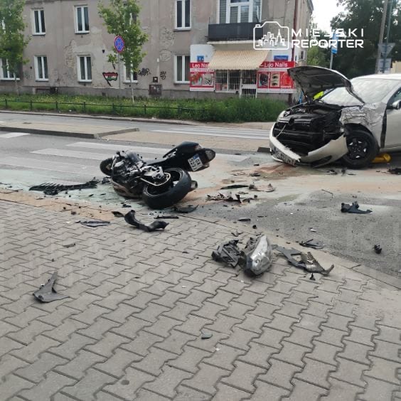 Zniszczony motocykl leży na jezdni obok rozbitego samochodu, w tle widoczny budynek mieszkalny.
