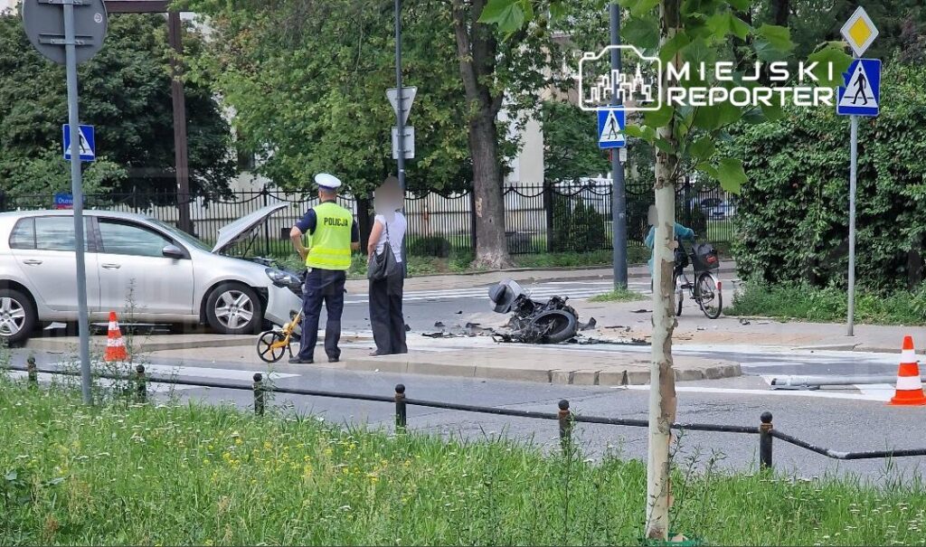 Policjant w kamizelce odblaskowej bada miejsce wypadku, obok uszkodzone auto i rozbite motocykl na skrzyżowaniu.