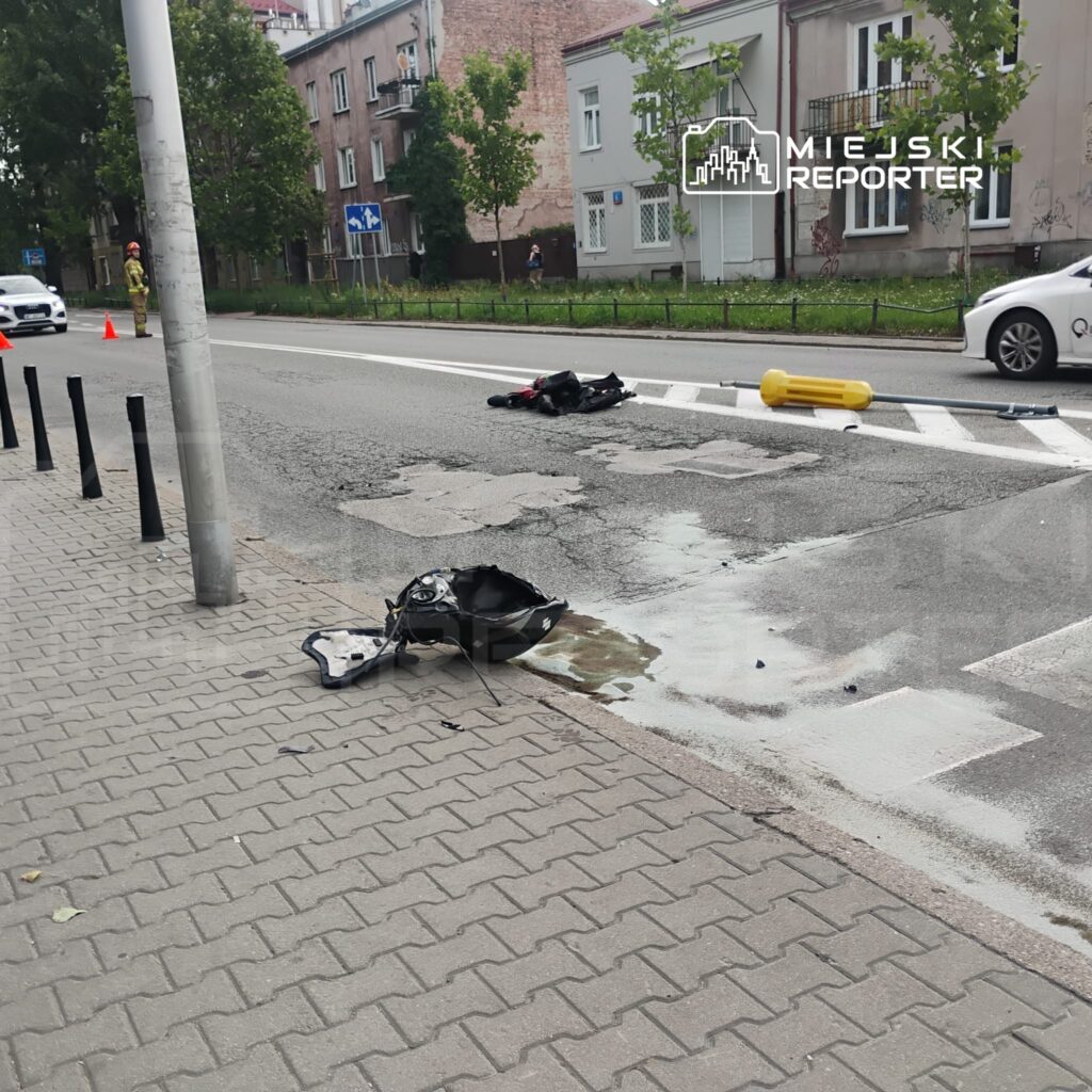 Na jezdni leży uszkodzona część motocykla, obok widoczna plama płynu. W tle samochód osobowy i budynki.