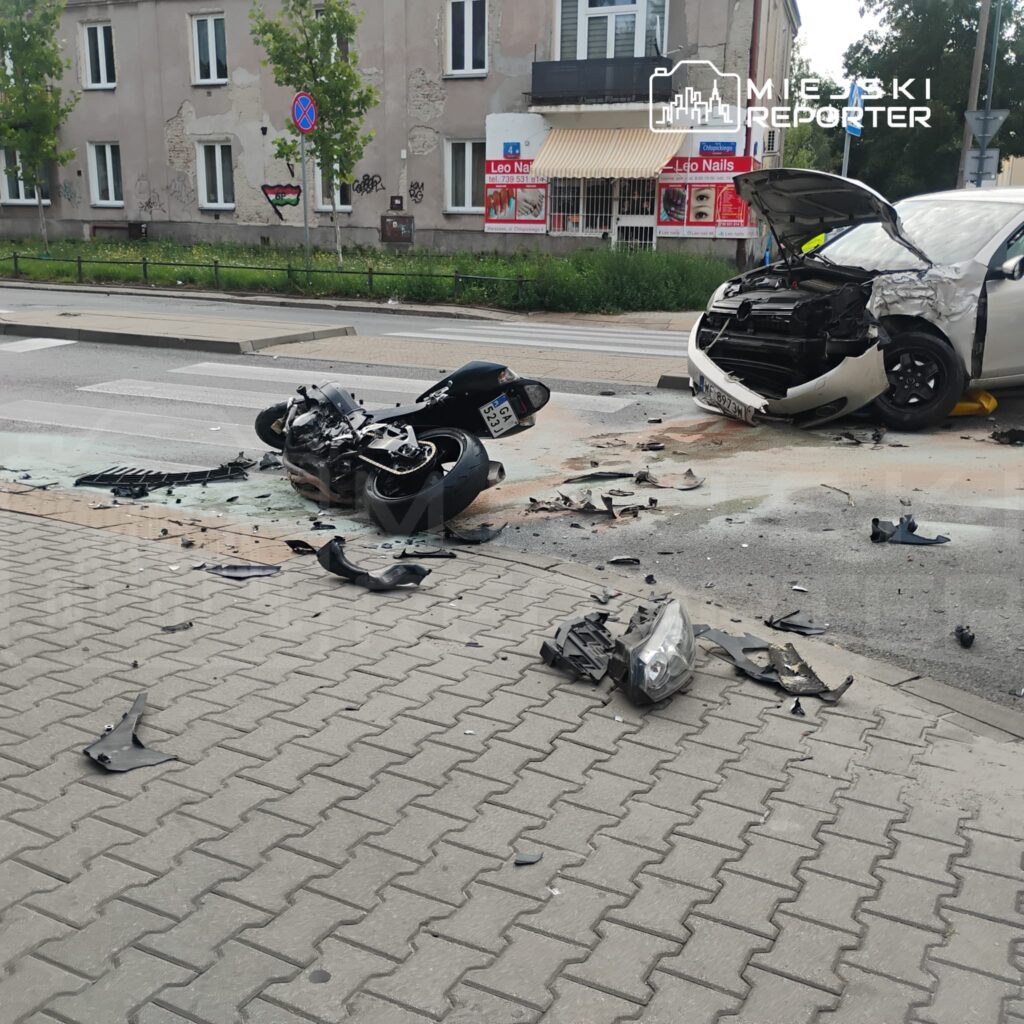 Wypadek drogowy z udziałem motocykla i osobowego samochodu, na jezdni leżą uszkodzone elementy pojazdów.